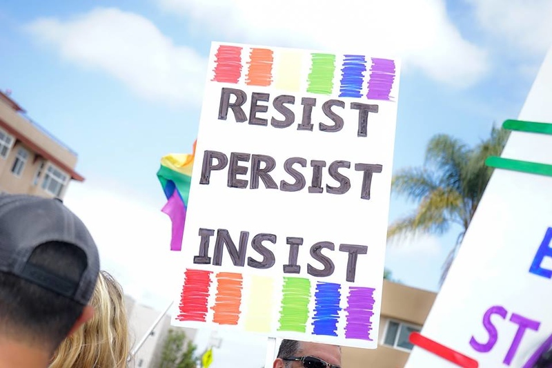 A protestor holds a sign that reads resist, persist, insist on a white background with rainbow bands